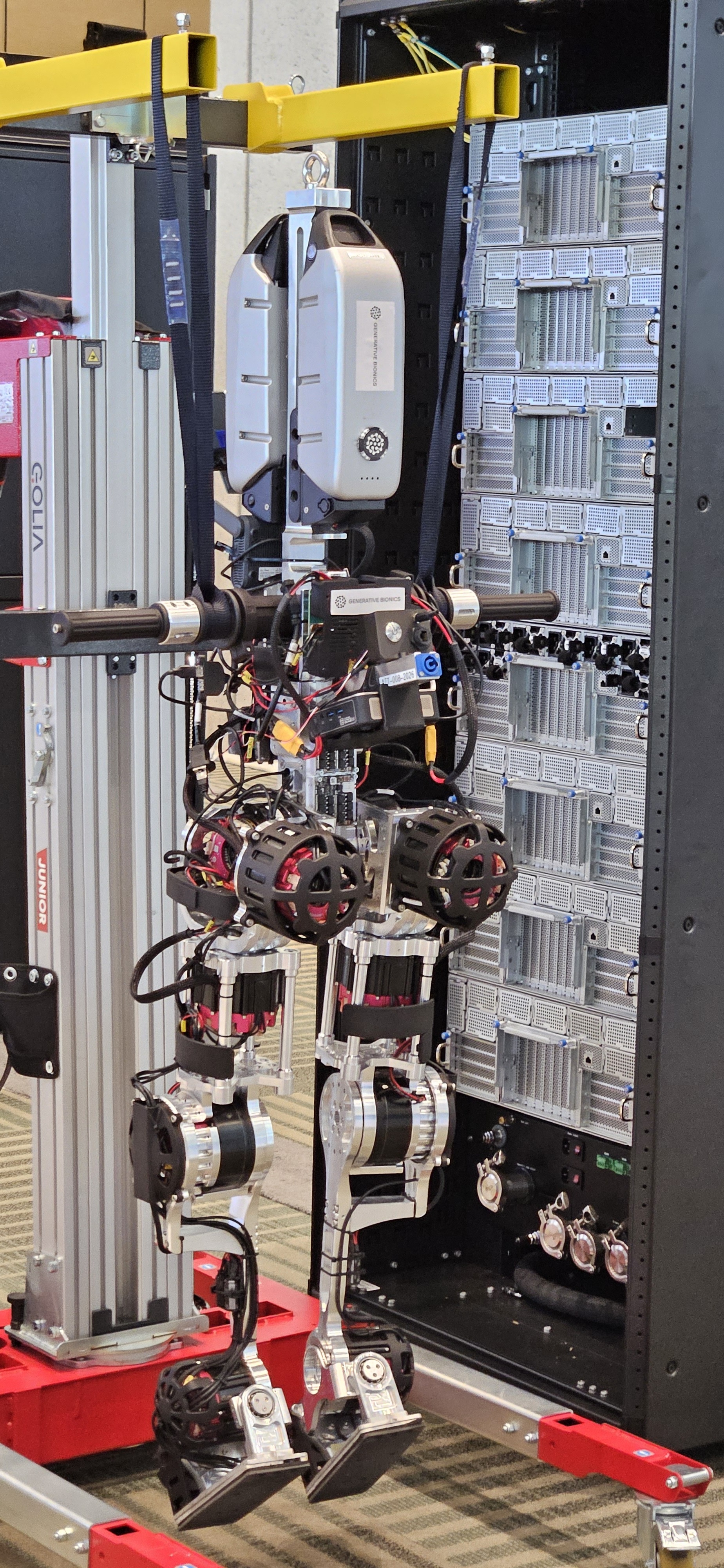 Close-up of the same Generative Bionics humanoid's hip and leg actuators, hanging from the support gantry, with a rack-mounted compute unit visible behind it.
