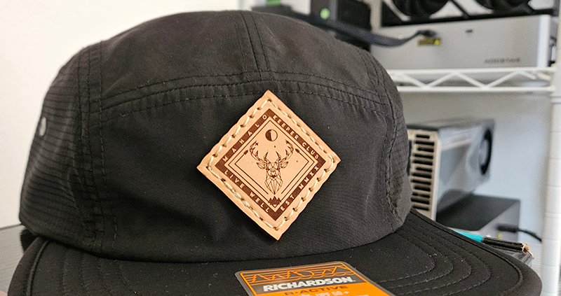 A black runner-style cap with a diamond-shaped leather patch reading Mahalo Keeper Club, Live Well, with a buck's head silhouette in the center.